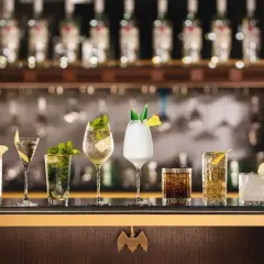 A row of seven colorful cocktails on a bar, with bottles in the background.